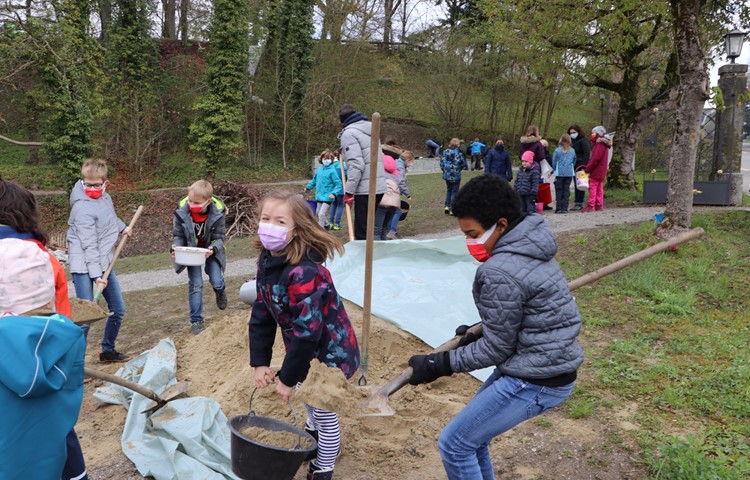 Viertklässler schaufeln lehmhaltigen Sand in ihre Eimer und tragen ihn zum Loch hinter der Trockenmauer (Bildmitte), wo er festgestampft wird. Viertklässler schaufeln lehmhaltigen Sand in ihre Eimer und tragen ihn zum Loch hinter der Trockenmauer (Bildmitte), wo er festgestampft wird.